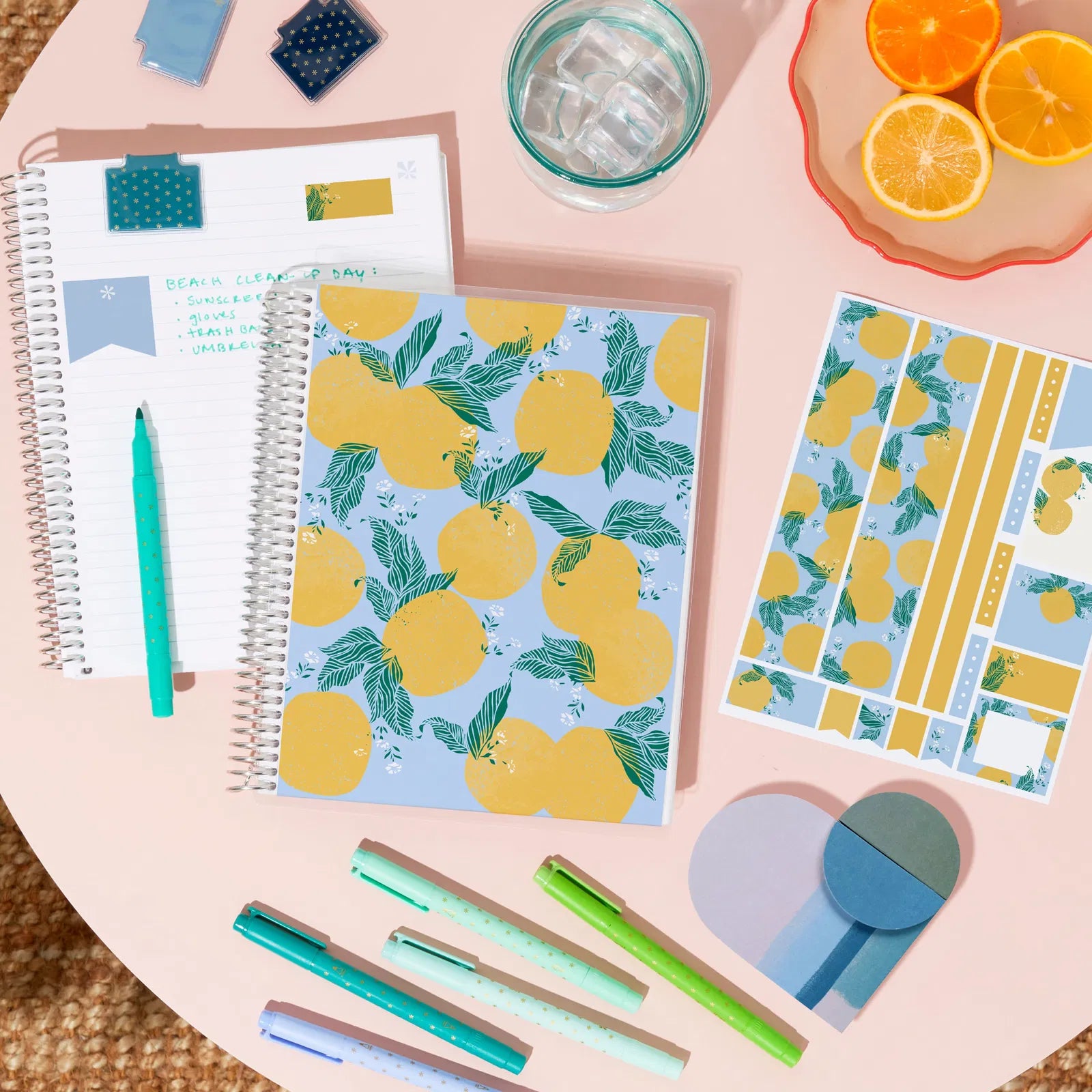 Notebook with lemon pattern, pens, and oranges on a pink surface