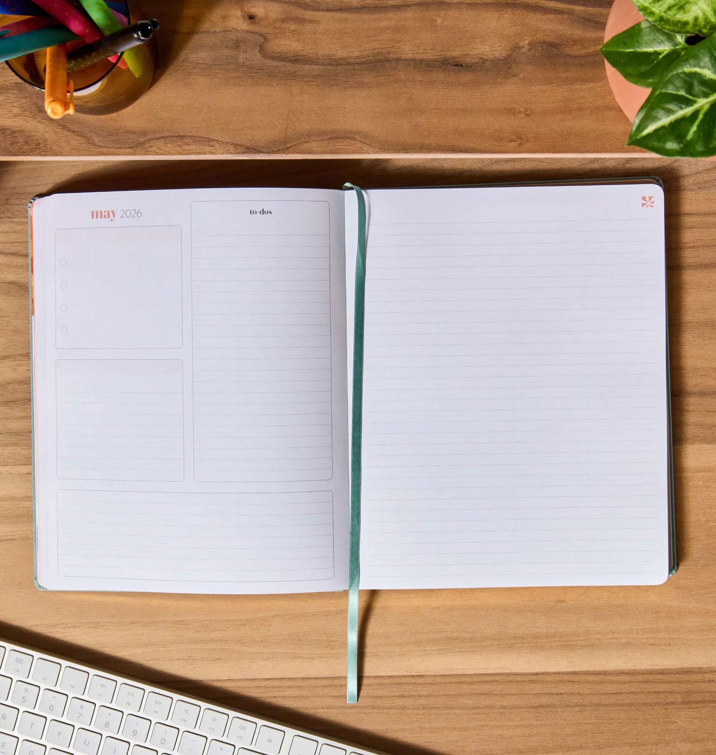 Open notebook on a wooden desk with a keyboard and plant in the background
